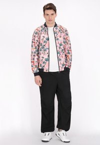 Roze bomberjack met tropische print, voorzien van donkerblauwe geribde manchetten en kraag, gecombineerd met zwarte broeken en witte sneakers.