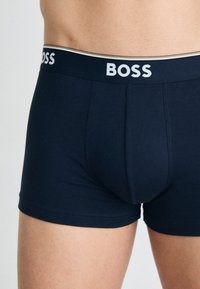 Tengeri kék pamut boxeralsó fehér "BOSS" derékpánttal, sima textúrával és testhezálló kialakítással. Nincsenek látható minták vagy díszítések.