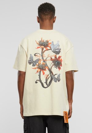 Cremfarbendes, übergroßes T-Shirt mit einem floralen und Schmetterlingsdesign auf dem Rücken, mit orangefarbenen Akzenten und einem seitlichen Etikett.