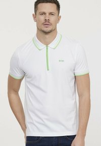 Witte polo met groene kraag en ritssluiting, met korte mouwen en een subtiel logo op de borst. Zachte, gladde stoftextuur.