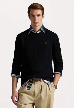 Zwarte gebreide trui met een ronde hals, gedragen over een blauw- en groen-geruit overhemd. Khaki broek en een bruine riem zijn zichtbaar.