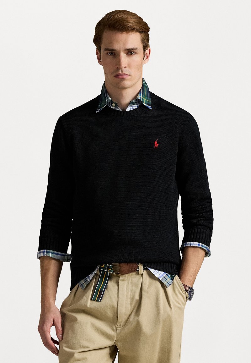 Suéter preto de malha com decote redondo, usado sobre uma camisa xadrez azul e verde. Calças cáqui e um cinto castanho são visíveis.