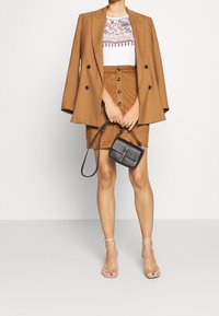 Brun dubbelknäppt kavaj över en vit mönstrad topp, kombinerad med en brun kjol med knappar framtill. Svart axelväska och nakna sandaletter med remmar.