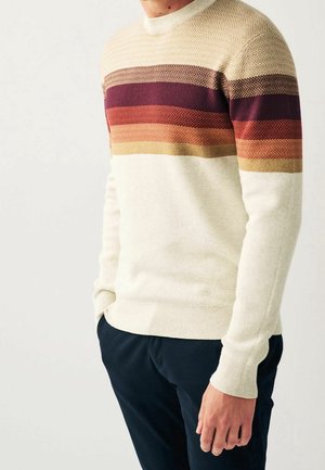 Mann trägt einen cremefarbenen Pullover mit horizontalen braunen, kastanienbraunen und orangen Streifen über der Brust und eine marineblaue Hose, steht mit den Händen in den Taschen.