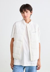 Jongere met kort donker haar draagt een witte gewatteerde vest over een wit T-shirt en blauwe spijkerbroek, staand tegen een plain witte achtergrond.