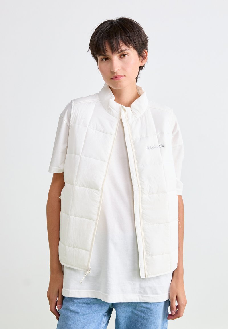 Jongere met kort donker haar draagt een witte gewatteerde vest over een wit T-shirt en blauwe spijkerbroek, staand tegen een plain witte achtergrond.