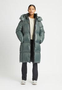 Dik, waterdichte parka in donkergroen met een capuchon, ritssluiting, zwarte accenten en gestikte panelen. Draagt over een lichte trui en broek.