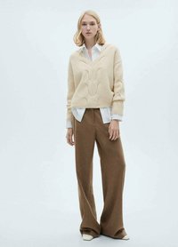 Beige gestrickter Pullover mit V-Ausschnitt und Zopfmuster über einem weißen Hemd getragen. Braune weite Hose und crèmefarbene Schuhe vervollständigen das Outfit.