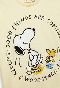 Jasnożółta koszulka z czarnymi i żółtymi ilustracjami Snoopy'ego i Woodstocka, z napisem "NADCHODZĄ DOBRE RZECZY" okrążającym je.