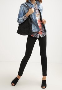 Veste en jean portée sur un chemisier à fleurs, associée à un jean skinny noir et des chaussures noires à bout ouvert. Porte un sac à main en tissu noir.