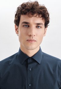 Ciemnoniebieska koszula zapinana na guziki z małym białym wzorem w kropki. Posiada klasyczny kołnierzyk i widoczne guziki z przodu.