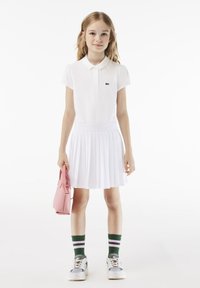 Biała koszulka polo z krótkimi rękawami i logo Lacoste, zestawiona z białą plisowaną spódnicą, zielono-różowymi skarpetkami w paski i szarymi trampkami.