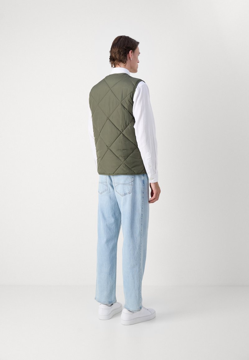 Gepolsterte olivgrüne Weste mit Rautennähten, die über einem weißen Hemd getragen wird. Hellblaue Jeans und weiße Sneakers runden das Outfit ab.