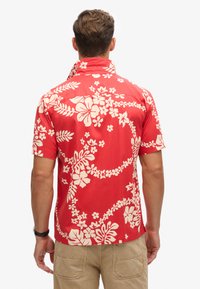 Camisa roja de manga corta con patrones florales blancos, que incluyen hibiscos y hojas, con un diseño en el cuello trasero y un ajuste relajado.