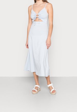 Femme portant une robe midi bleu clair à fines bretelles, découpe sur le devant, détail froncé avec lien et sandales compensées blanches, debout sur un sol gris.