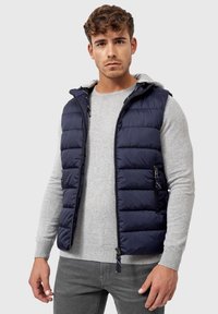Marineblå puffervest med lynlås fortil, sidelommer og en grå hætte. Båret over en lys grå sweater, der viser en quiltet tekstur.