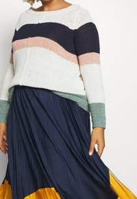 Sweter w paski w kolorze kremowym, granatowym, różowym i zielonym, w połączeniu z lejącą, granatową spódnicą plisowaną z żółtym wykończeniem. Widoczne detale tekstury materiału.