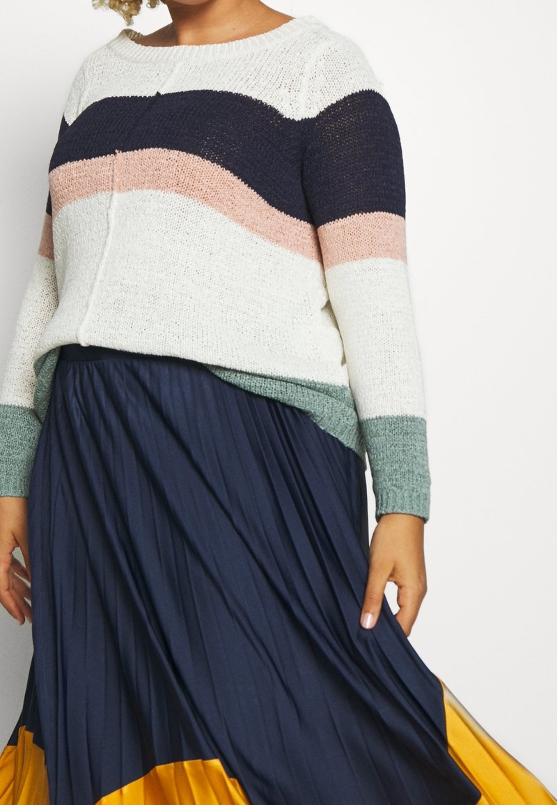 Sweter w paski w kolorze kremowym, granatowym, różowym i zielonym, w połączeniu z lejącą, granatową spódnicą plisowaną z żółtym wykończeniem. Widoczne detale tekstury materiału.