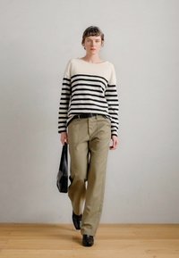 Ριχτή μπλούζα με μαύρες και λευκές ρίγες, σε χαλαρή γραμμή, συνδυασμένη με ελαιόχρωμες παντελόνες και μαύρα παπούτσια. Το μοντέλο κρατά μια μαύρη τσάντα.
