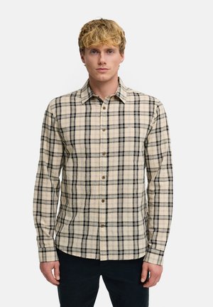 Młody mężczyzna o blond włosach, ubrany w beżowo-czartą koszulę w kratę zapinaną na guziki oraz ciemne spodnie, stojący na jednolitym białym tle.