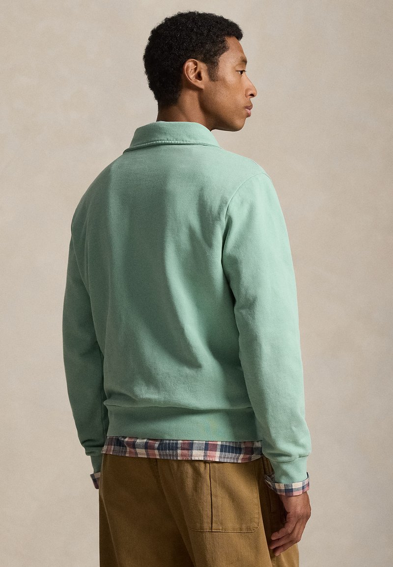 Man facing sideways wearing light green sweatshirt over plaid shirt and brown pants against neutral background.