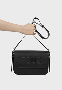 Czarna skórzana torba na ramię z wytłoczonym logo "TOUS", prostokątny kształt, regulowany pasek i srebrne akcenty metalowe.