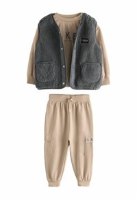 Dvoudílný outfit složený z šedé, huňaté vesty se cvoky a dvěma kapsami, spárovaný s béžovými jogger kalhotami s elastickým pasem a postranními kapsami.
