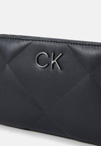 Melna ādas rokassomiņa ar quilted dizainu, reljefu "CK" logo un gludu tekstūru. Šūti maliņas uzlabo izturību.