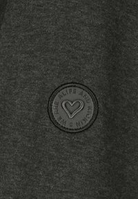 Dark gray fabric featuring a round black rubber patch with a heart design and the text "WE ARE ALIFE AND KICKIN" embossed around the edge.