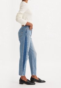 Jeans de tiro alto azul claro con paneles laterales en contraste, combinados con un suéter de punto blanco y mocasines negros, luciendo con las manos en las caderas.