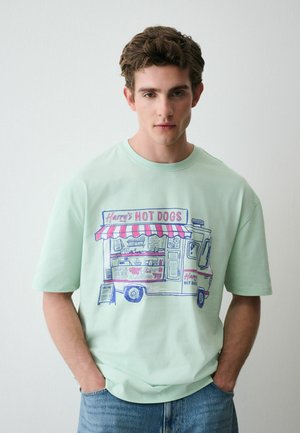 Junger Mann trägt ein hellgrünes T-Shirt mit einem bunten, illustrierten Hotdog-Foodtruck und der Aufschrift „Harry's Hot Dogs“ und steht vor einer schlichten Wand.