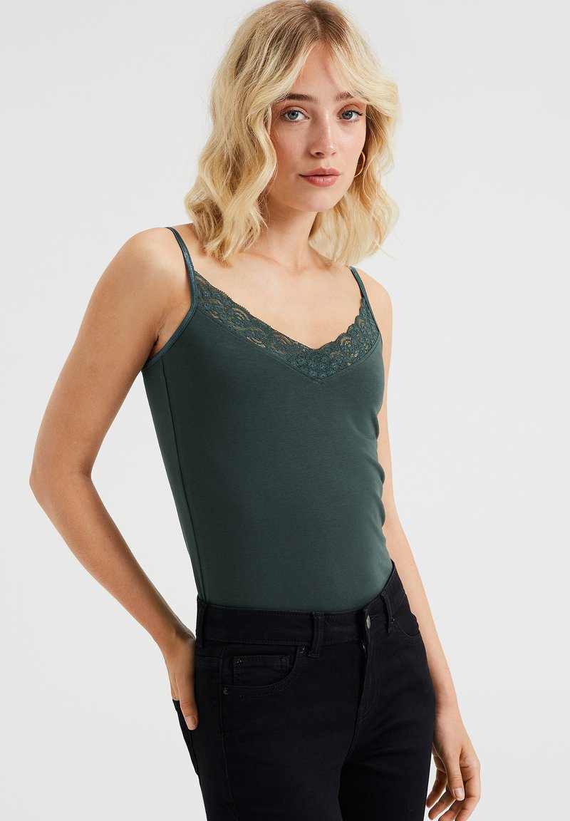 Grön cami med spetsdetaljer, figursydd design och tunna band, bärs med svarta jeans. Slät textur, v-ringning och delikat spets.