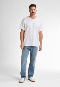 Tricou alb din bumbac cu guler rotund, având un text grafic albastru. Asortat cu blugi deschiși la culoare și adidași albi.