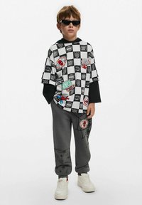 Kortärmad, rutmönstrad svartvit skjorta med olika grafiska lappar; grå sweatpants med spindelmönster, kombinerat med vita sneakers.