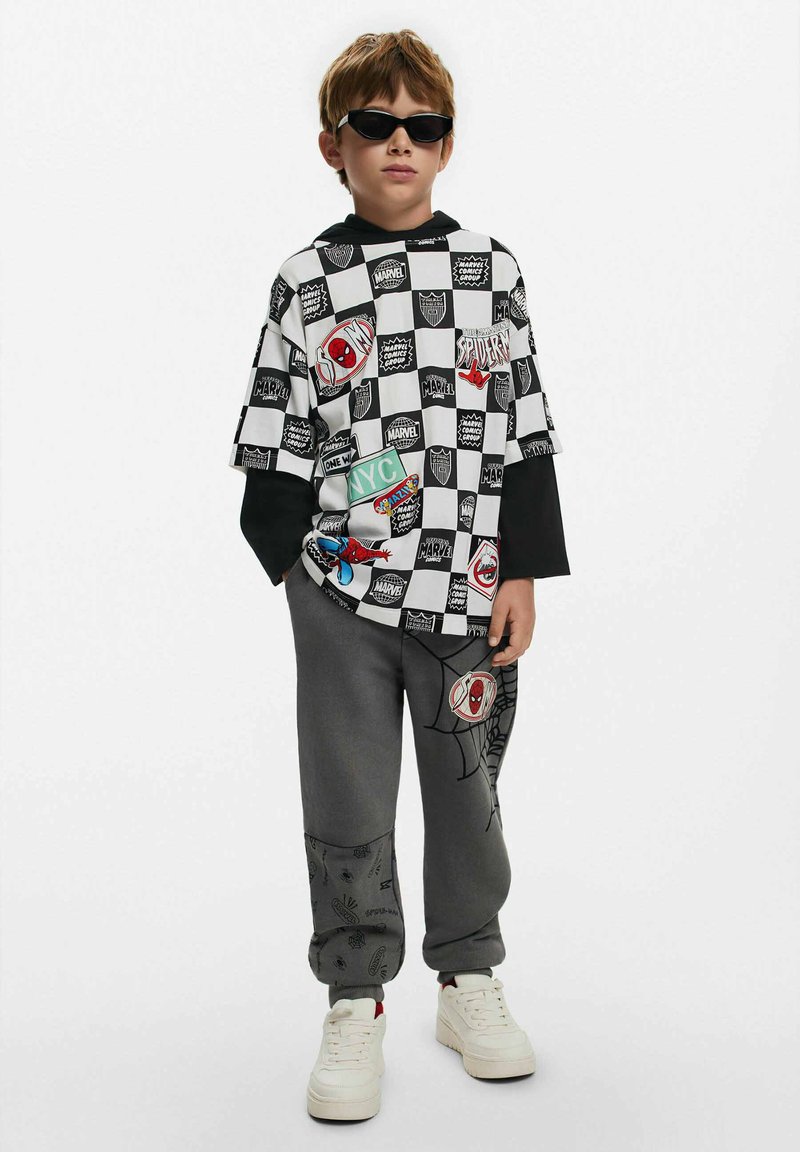 Kortärmad, rutmönstrad svartvit skjorta med olika grafiska lappar; grå sweatpants med spindelmönster, kombinerat med vita sneakers.