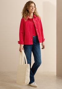 Ljusröd jacka med knäppning, jeans i denim och vita sneakers. Håller en krämfärgad, vadderad tote-väska.
