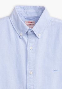 Lyseblå skjorte med knapper, spids krave, hvide knapper og et broderet Levi's-logo i matchende blå på venstre side.