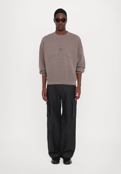 Sweat-shirt gris à coupe ample, épaules tombantes et impression textuelle. Pantalons cargo noirs avec plusieurs poches. Le modèle porte des lunettes de soleil et des chaussures noires.