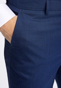 Marineblaue maßgeschneiderte Hose aus strukturiertem Stoff, mit einer Seitentasche und glattem Bund. Nahaufnahme des Stoffdetails und der Handplatzierung.