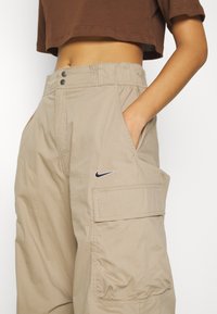 Pantalones cargo beige con cinturilla elástica, bolsillos laterales y un pequeño logo negro de Nike en el frente. Textura de tela suave.