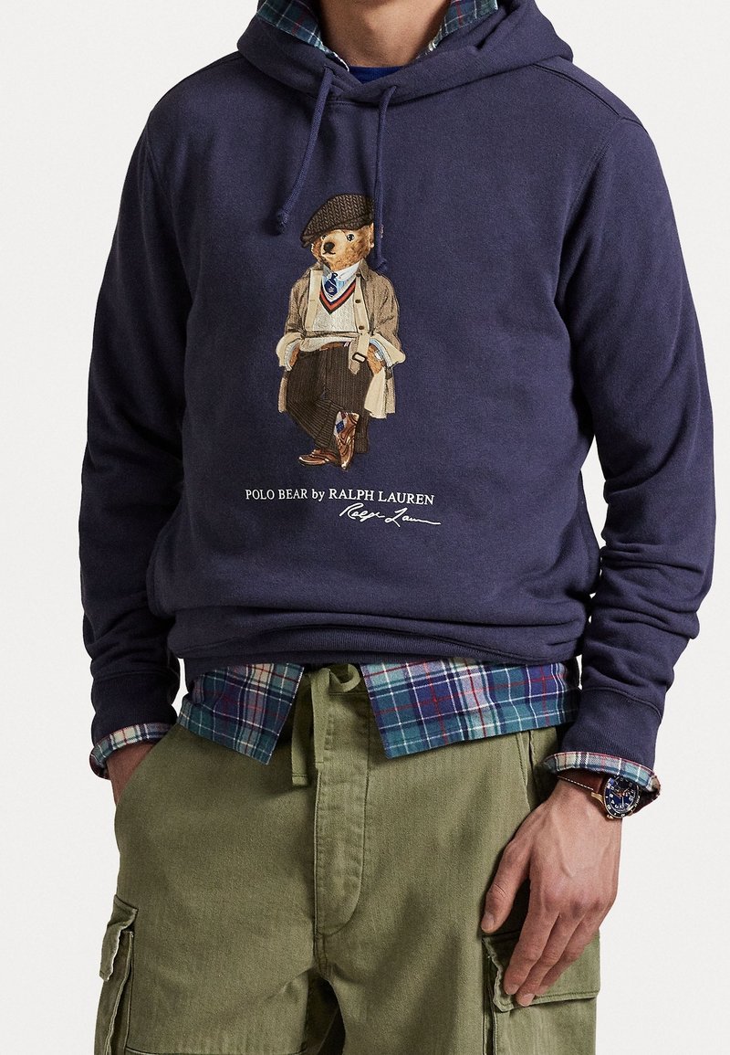 Personne portant un sweat à capuche bleu marine avec un motif Polo Bear, une chemise à carreaux en dessous, un pantalon cargo olive et une montre au poignet gauche.