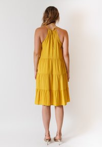 Abito giallo senza maniche con design a strati e scollatura con nastro legato sulla schiena; la texture morbida e il tessuto fluido creano una silhouette comoda.