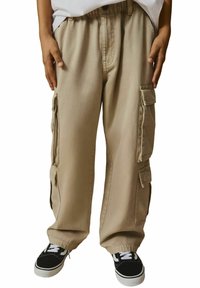Un pantalon cargo beige présente plusieurs poches latérales, une coupe décontractée et une taille à cordon, associé à des baskets noires avec des lacets blancs.