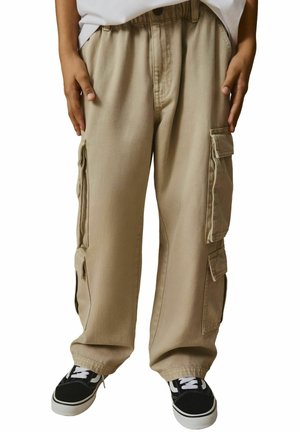 I pantaloni cargo beige presentano numerosi taschini laterali, una vestibilità comoda e una vita con cordino, abbinati a sneakers nere con lacci bianchi.