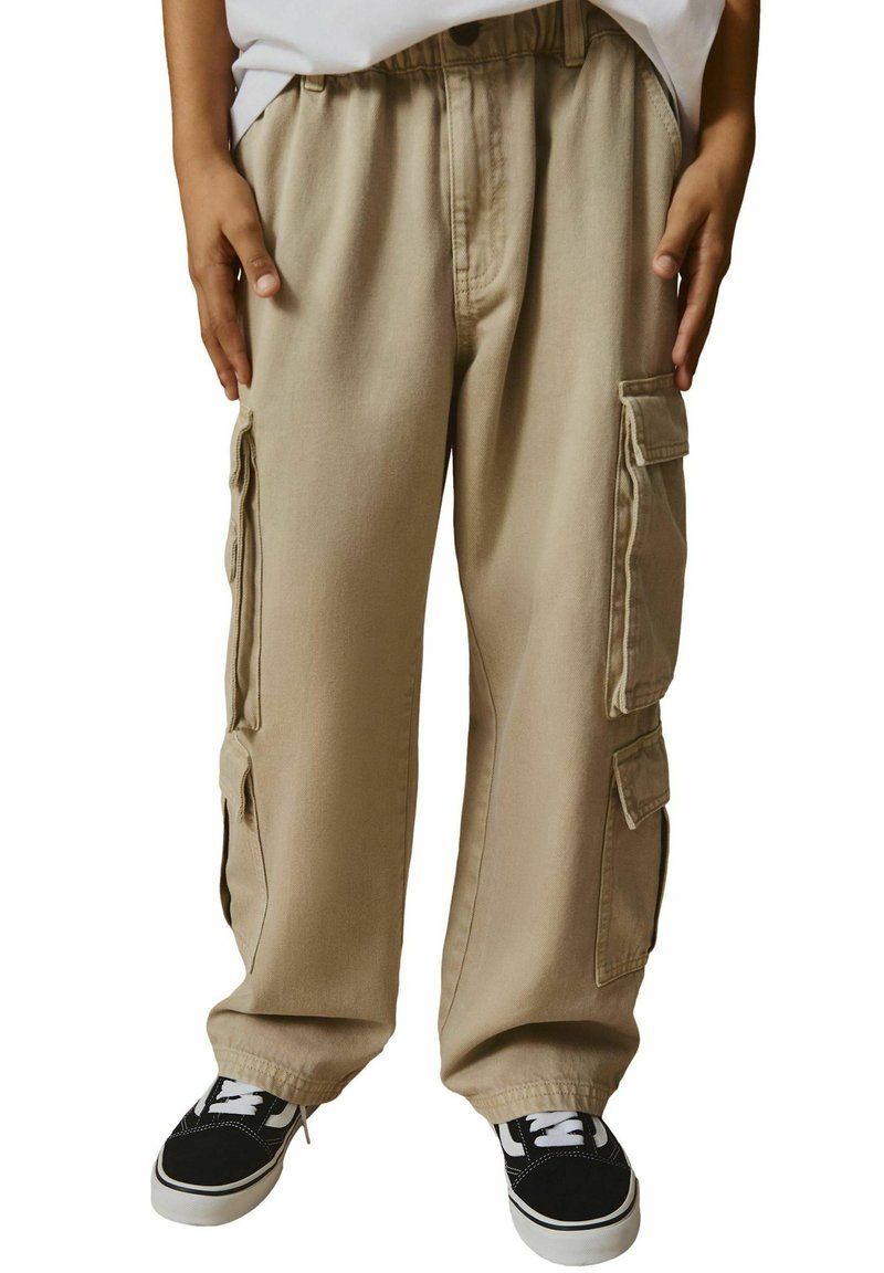 Un pantalon cargo beige présente plusieurs poches latérales, une coupe décontractée et une taille à cordon, associé à des baskets noires avec des lacets blancs.