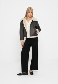 Brun lærjakke med en kremfarget faux fur-krage og mansjetter, glidelås foran, brukt over en lys skjorte og parret med svarte vide bukser.