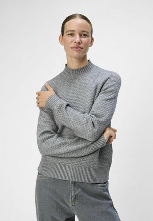 Suéter de punto gris claro con un patrón texturizado, cuello alto y puños acanalados. Combinado con jeans grises, usado por una persona con las manos entrelazadas.
