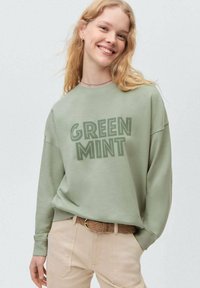 Mintgroene sweatshirt met de tekst "GREEN MINT", ontspannen pasvorm en lange mouwen, gecombineerd met lichtbeige broek en een gestructureerde riem.