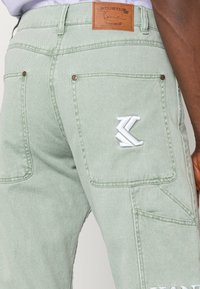 Light green denim cargo pants featuring rear pockets, embroidered white logo detail, and a brown leather patch at the back waist.