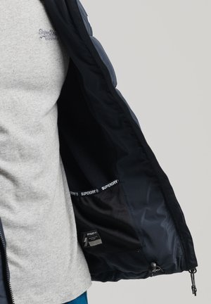 En person iført en grå sweatshirt åbner en marineblå Superdry-jakke, der viser det sorte indvendige for og den mærkede inderlomme.